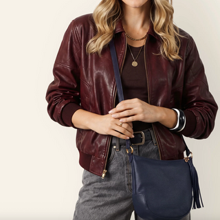 A woman wearing a burgundy faux-leather bomber jacket, dark brown top, gray jeans, and carrying a navy blue pebbled leather crossbody bag with gold-tone hardware.