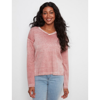 A woman with long, wavy black hair is wearing a pink, heathered, long-sleeve V-neck top with light blue jeans, standing against a light gray studio background.