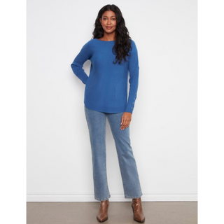 A woman modeling a blue long-sleeve knit top with light-wash blue jeans and brown ankle boots, standing against a plain white wall.