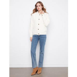 A woman modeling a cream-colored knit button-up jacket with silver buttons, paired with blue jeans and tan ankle boots, standing against a white wall.