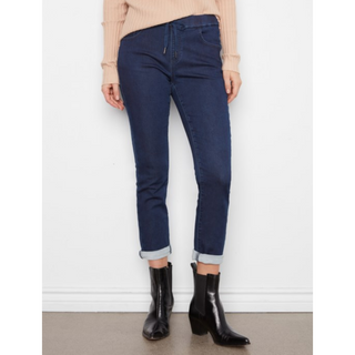 A person wearing dark blue skinny jeans with rolled cuffs, paired with a beige ribbed sweater and black ankle boots, standing against a white wall.