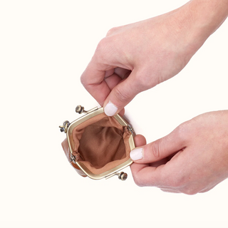 Close-up of a person's hands holding open a small kiss-lock style clutch bag with a tan interior lining and gold-tone metal frame, on a plain white background.