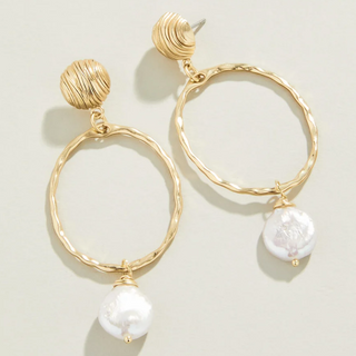 A pair of gold-toned dangle earrings with textured circular hoops, wrapped spherical tops, and white baroque pearl drops, shown on a light beige background.