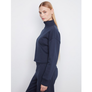 A woman modeling a navy blue ribbed knit turtleneck sweater and matching pants against a light gray studio background.