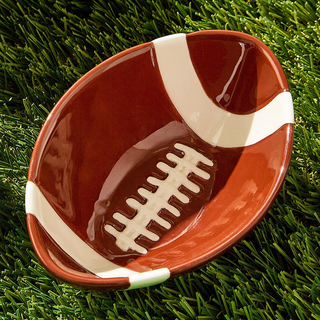 A football-shaped ceramic dish with a glossy brown body, white stripes, and white laces, photographed on green artificial turf.