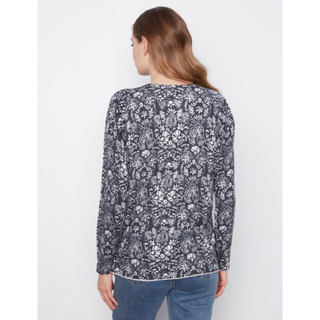 A woman viewed from the back wearing a long-sleeve sweater with an all-over white floral and paisley print on a dark navy or charcoal base, paired with blue jeans against a plain light gray studio background.