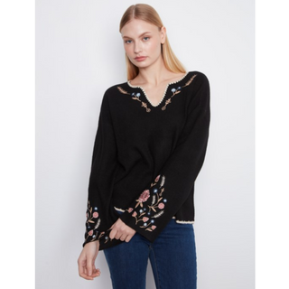 A woman modeling a black long-sleeve knit top with colorful floral embroidery, paired with blue jeans against a light gray studio background.