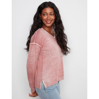 A woman with long, wavy dark hair is shown from the side/back wearing a heathered dusty pink knit sweater with white contrast stitching details, paired with light blue jeans, against a plain light gray studio background.