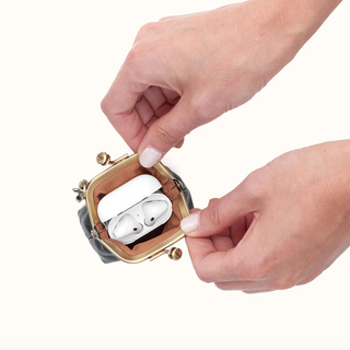 A close-up of a person's hands holding an open small metallic clutch purse, showing a white wireless earbud charging case inside, on a plain white background.