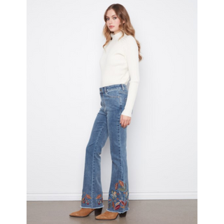 A woman modeling a pair of blue denim flare jeans with colorful embroidered details near the hem, paired with a white mock-neck sweater and brown ankle boots, standing against a white wall.