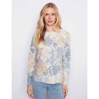 A woman modeling a long-sleeve knit sweater with a beige and light blue abstract botanical pattern, paired with light-wash blue jeans, against a plain light gray studio background.
