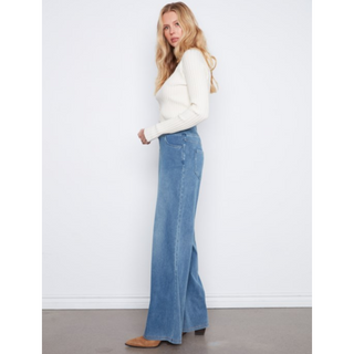 A woman stands in profile wearing a cream ribbed knit sweater and high-waisted, wide-leg blue jeans against a plain white wall.