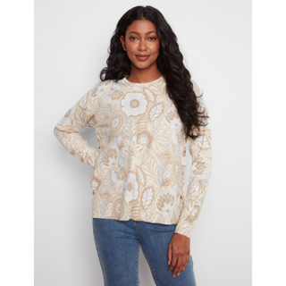 A woman modeling a cream and beige floral knit sweater with blue jeans against a light gray studio background.