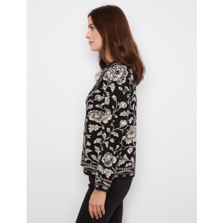 A woman shown in side profile wearing a long-sleeve sweater with a black and white floral and vine pattern, paired with black pants, against a plain light gray studio background.