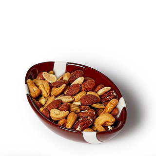 An oval ceramic bowl with a glossy dark red interior and white side accents, filled with mixed nuts including cashews, almonds, and pecans, photographed on a clean white background.