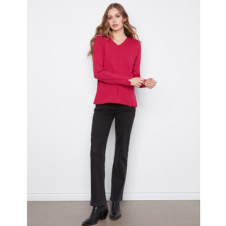 A woman modeling a bright pink long-sleeve V-neck sweater paired with black pants and black boots, standing against a plain white wall.