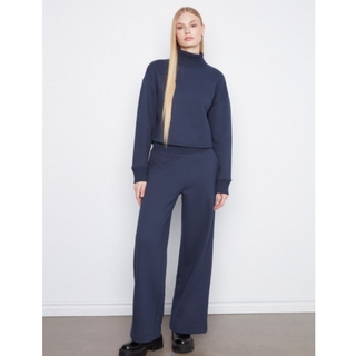 A model stands against a white wall wearing a matching navy blue knit set consisting of a cropped mock-neck long-sleeve top and wide-leg trousers, paired with black platform shoes.