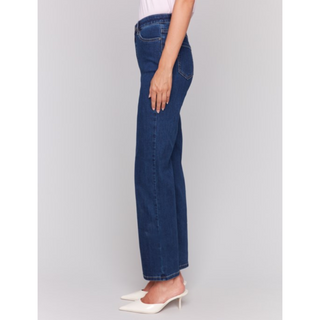 A side profile view of a model wearing dark blue wide-leg jeans and white pointed-toe mule heels against a light gray studio background.