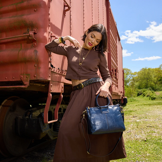 A woman posing outdoors next to a red train car, wearing a brown long-sleeve crop top and matching skirt, holding a navy blue leather handbag.