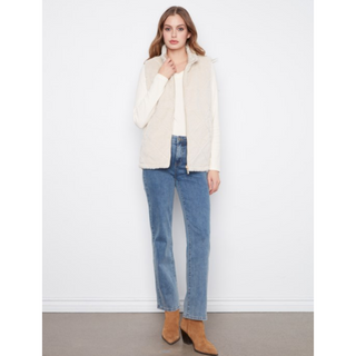 A woman stands against a plain white wall wearing a cream-colored fuzzy sleeveless vest over a white top, blue jeans, and tan ankle boots.