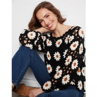 A woman wearing a black crochet knit sweater with a white and brown daisy pattern, paired with blue jeans and brown boots, sitting against a light gray wall.