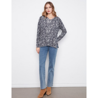 A woman modeling a long-sleeve V-neck top with a gray and white floral pattern, paired with blue jeans and brown ankle boots, standing against a white wall.