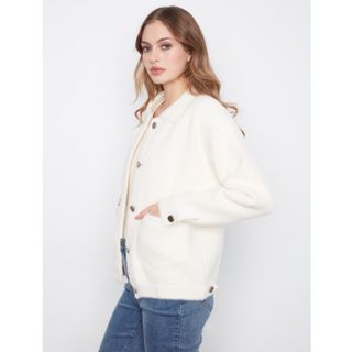A woman wearing a cream-colored, fuzzy button-up jacket with dark buttons and blue jeans, standing against a plain light gray background.