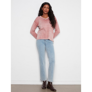 A woman modeling a pink long-sleeve sweater with a V-neck, paired with light blue jeans and dark brown boots, standing against a plain white wall.