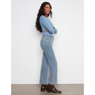 A woman modeling a light blue long-sleeve top and light-wash blue straight jeans with brown leather ankle boots, standing against a white wall.