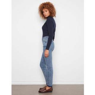 A woman with curly light brown hair stands in profile wearing a dark navy ribbed long-sleeve top, light blue skinny jeans, and brown leather loafers against a plain white wall.