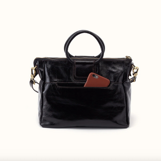 A black leather handbag with a top handle, gold-tone hardware, and a brown leather accent on the front pocket, shown against a plain white background with a smartphone tucked into the pocket.