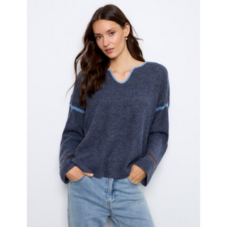 A woman modeling a navy blue knit sweater with light blue trim details on the v-neck and sleeve cuffs, paired with light-wash blue jeans against a plain light gray background.