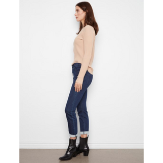 A woman stands in profile wearing a beige ribbed long-sleeve top, dark blue cropped jeans, and black ankle boots against a plain white wall.