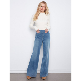 A woman modeling a white ribbed mock-neck sweater and high-waisted, wide-leg blue denim jeans against a plain white wall.