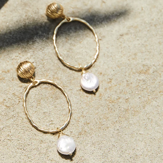 A pair of gold-toned dangle earrings with textured circular hoops, small ribbed bead tops, and white baroque pearl drops, photographed on a sandy beige surface with natural shadows.