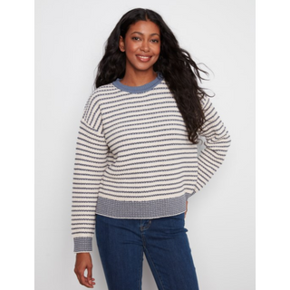 A woman wearing a cream and navy striped knit sweater with blue trim, paired with dark blue jeans, standing against a light gray studio background.