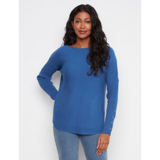 A woman modeling a blue long-sleeve knit sweater with a boat neckline and decorative silver buttons on one sleeve, paired with light blue jeans against a plain light gray background.