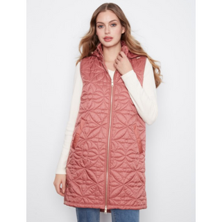 A woman modeling a sleeveless, quilted, zip-front puffer vest in a dusty rose pink color, worn over a cream long-sleeve top and blue jeans against a light gray studio background.