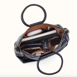 An open black leather handbag with gold-tone hardware, shown from above on a white background. The bag is filled with accessories, including a white oval case, a silver rectangular item, a black cardholder, and a pair of sunglasses.
