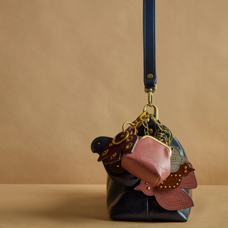 A dark navy leather bucket bag with a long strap, decorated with multiple layered leather flower charms in dusty pink, mauve, and brown, accented with gold-tone studs and rings, set against a warm tan background.