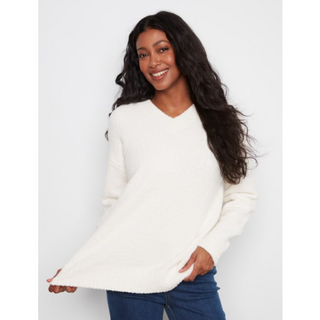A woman with long, wavy dark hair wearing a fuzzy off-white long-sleeve V-neck sweater and blue jeans, posing against a light gray studio background.