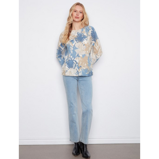 A woman stands against a plain white wall wearing a loose long-sleeve top with a blue and beige botanical pattern, light-wash jeans, and black ankle boots.