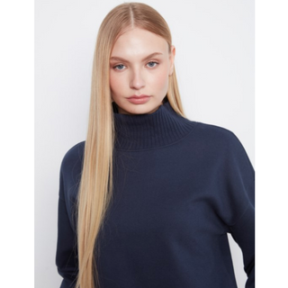 A blonde model wearing a navy blue ribbed turtleneck sweater, photographed against a light gray studio background.