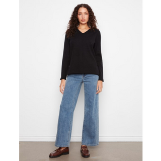 A woman modeling a black long-sleeve V-neck sweater and wide-leg blue jeans with brown leather loafers, standing against a plain white wall.