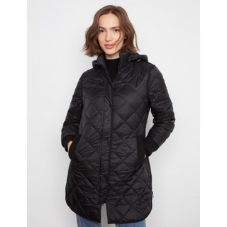 A woman wearing a black quilted hooded puffer coat over a black top and blue jeans, standing against a light gray studio background.