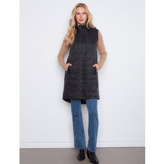 A woman stands against a white wall wearing a sleeveless black quilted puffer vest over a tan long-sleeve top, blue jeans, and black boots.