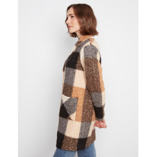 A woman shown in side profile wearing a long, fuzzy knit cardigan with a large patchwork plaid pattern in brown, beige, black, and gray, paired with blue jeans against a light gray studio background.
