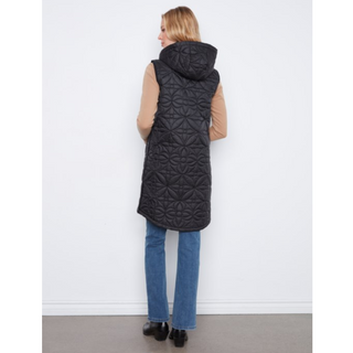 A woman viewed from the back wearing a long black quilted hooded puffer vest over a beige long-sleeve top and blue jeans, standing against a plain white wall.