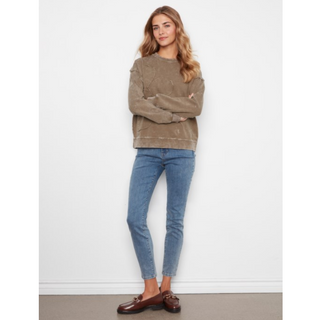 A woman stands against a white wall wearing a faded taupe-brown crewneck sweatshirt, blue skinny jeans, and brown leather loafers.