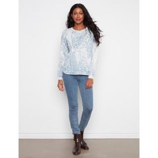 A woman modeling a light blue and white patterned long-sleeve knit top with blue jeans and dark brown ankle boots, standing against a plain white wall.